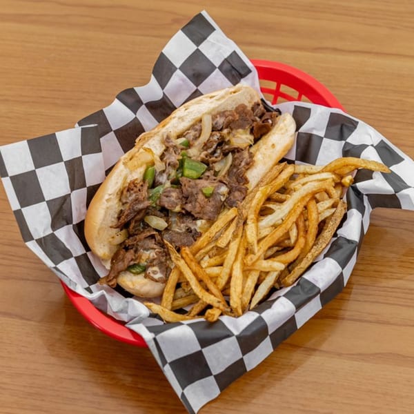 Cheesesteak sandwich with onions and peppers, served with fries in a red basket with checkered paper.
