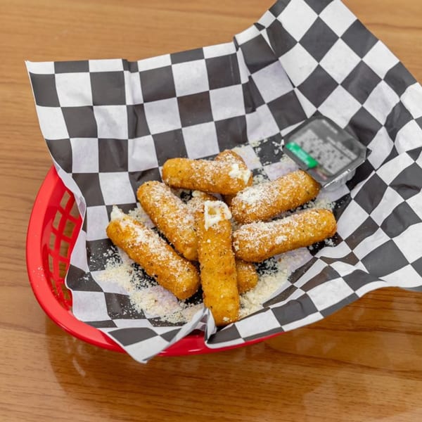 Golden-brown mozzarella sticks, sprinkled with parmesan, in a red basket with checkered paper and dipping sauce.